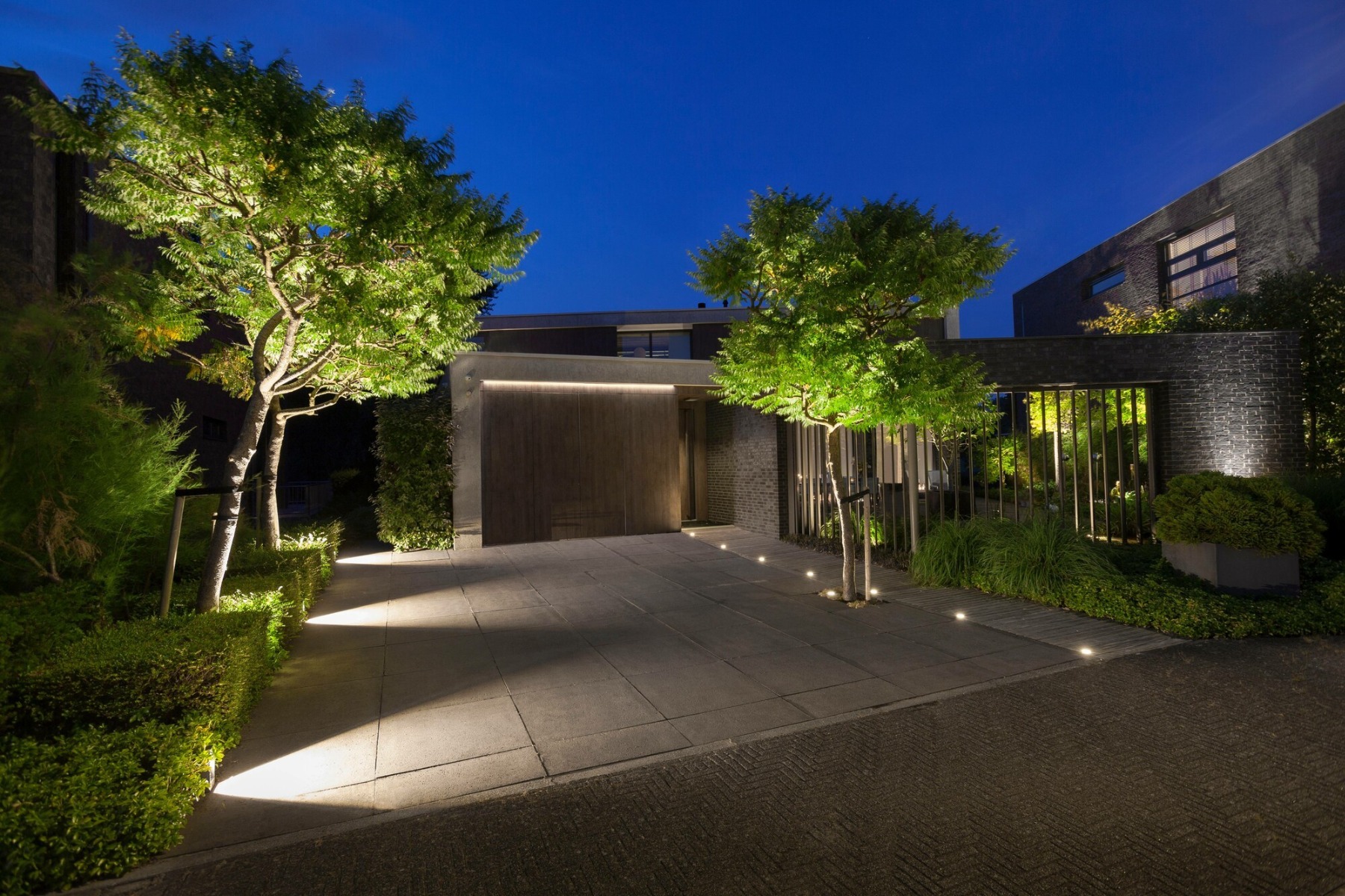 Eine Einfahrt mit Bäumen wird von zwei Seiten stimmungsvoll beleuchtet – auf der linken Seite werfen Stehleuchten breite Lichtkegel auf das Pflaster, auf der rechten Seite dienen Bodeneinbauleuchten als Hinweisbeleuchtung.