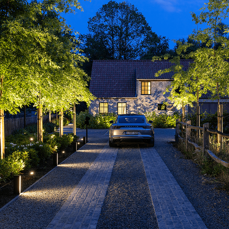 Eine effektvoll beleuchtete Einfahrt mit einem geparkten Auto. Wegeleuchten dienen als direkte Einfahrt-Beleuchtung, daneben sorgen angestrahlte Bäume für eine indirekte Beleuchtung der Einfahrt.