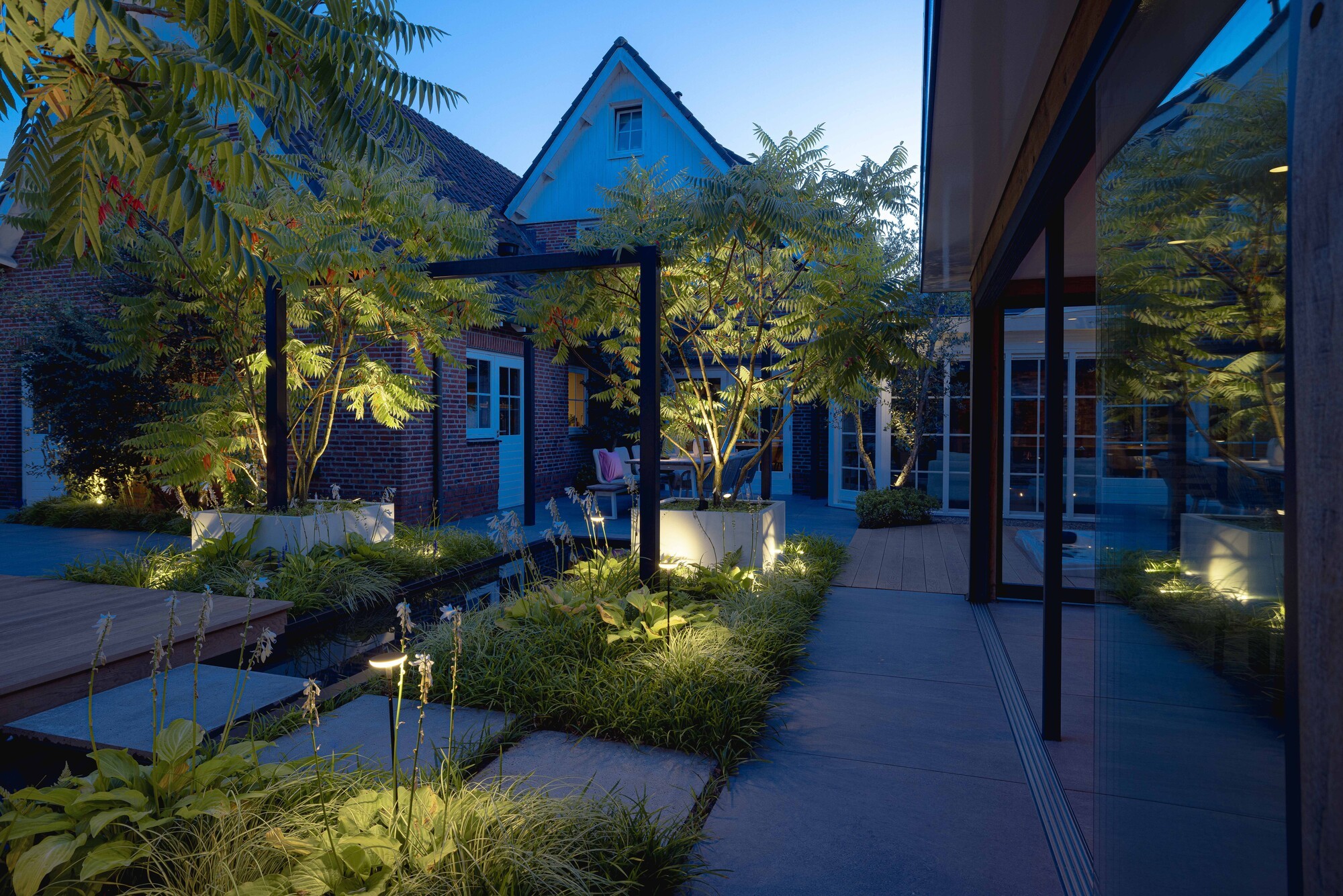 Ein Kabelverbinder, der zum Zubehör für in-lites Außenbeleuchtung gehört, ermöglicht eine einfache Installation nach dem Plug-and-Play-Prinzip und schließt zwei Kabel aneinander an. Im Hintergrund sind Blumen und eine Leuchte zu sehen.