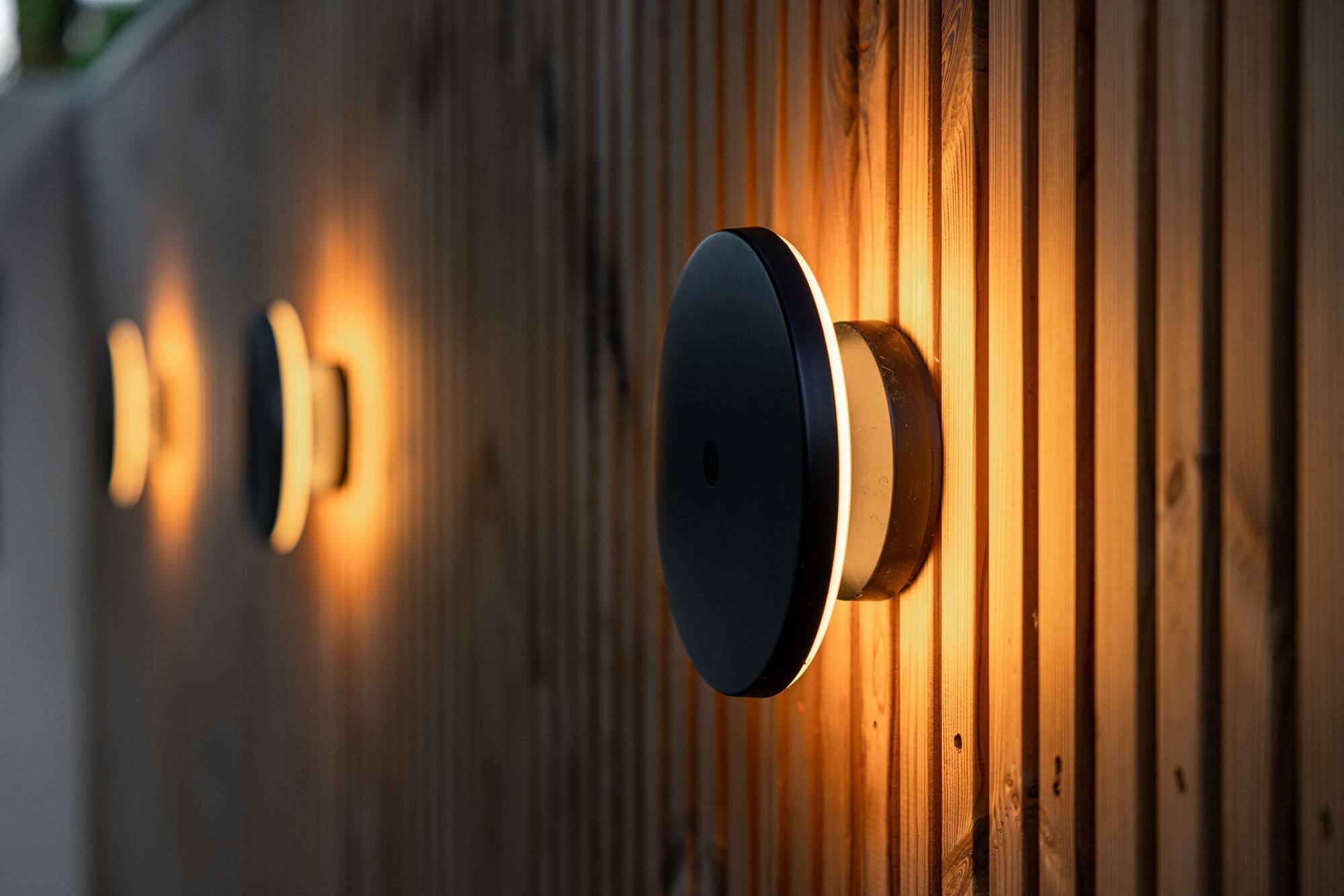 Drei runde Außenwandlampen tauchen eine Holzfassade in stimmungsvolles Licht.