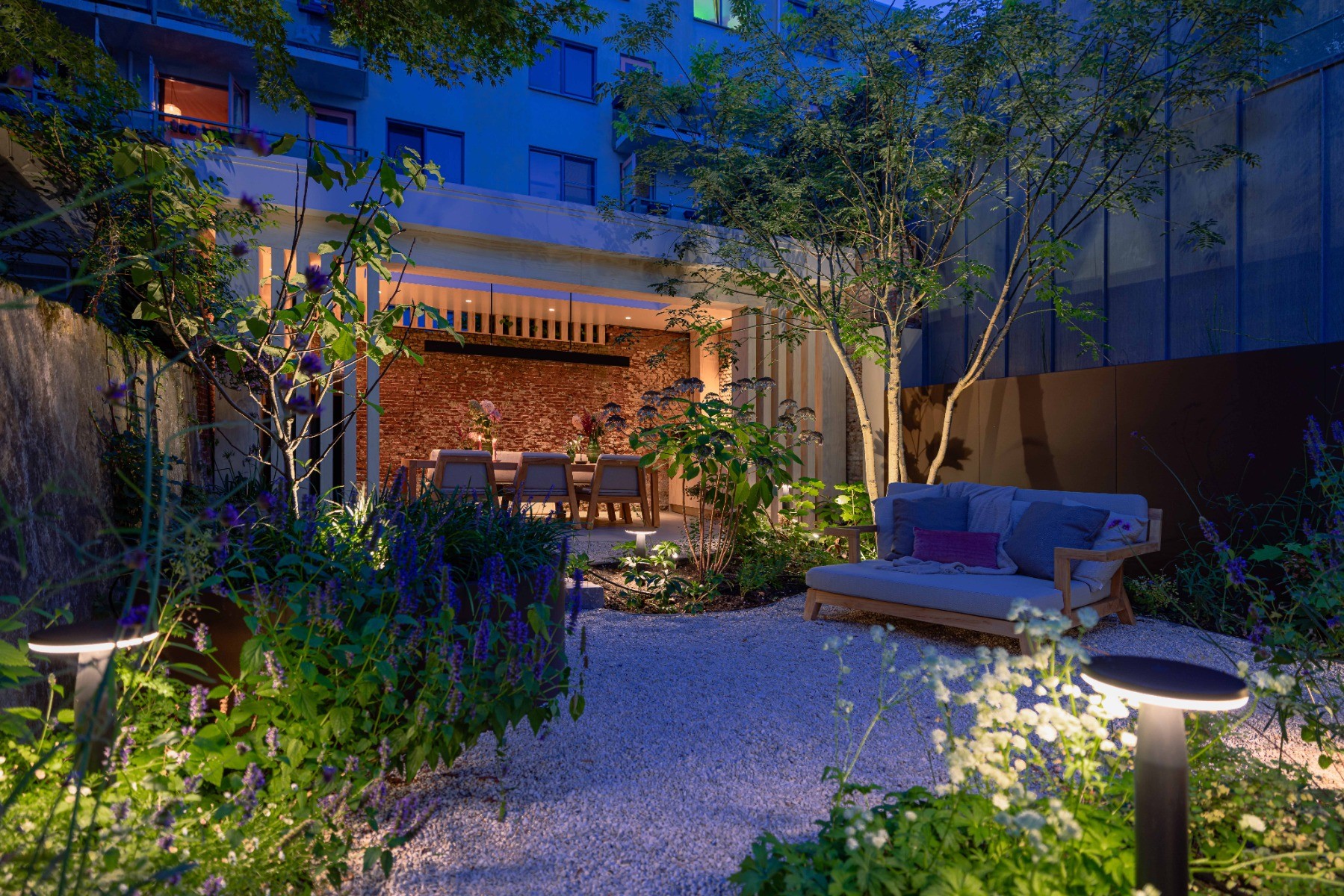 Ein moderner Garten mit Bepflanzung, einer gem&uuml;tlichen Sitzecke und einem Pool wird von verschiedenen Au&szlig;enleuchten erhellt. Im Mittelpunkt stehen die Zaun-, Baum- und Beetbeleuchtung.