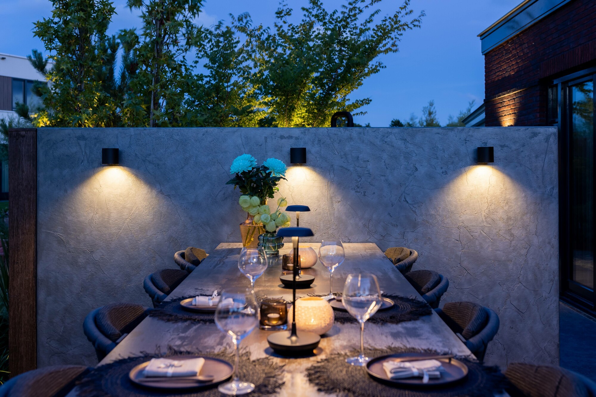 Eine Mauer mit drei LED-Außenwandleuchten, die eine angenehme Beleuchtung erzeugen. Davor ein gedeckter Gartentisch, der indirekt von den Außenwandleuchten und direkt von Tischleuchten erhellt wird. 