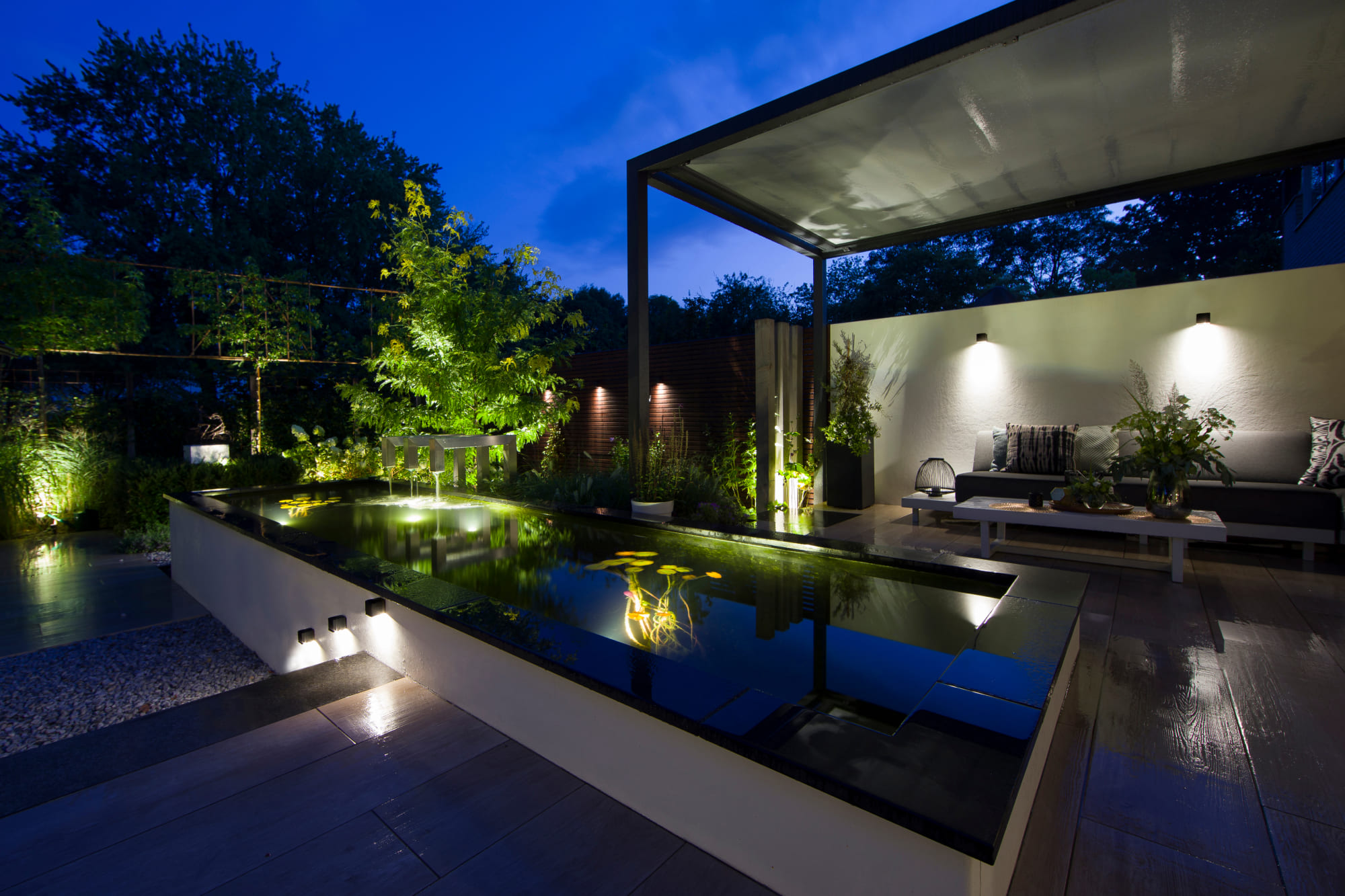 Eine elegante Außenanlage wird von diversen Außenleuchten erhellt. Im Zentrum sieht man einen modernen Teich, dessen Wasserpflanzen mit Unterwasserleuchten in Szene gesetzt werden. In der angrenzenden Sitzecke und entlang der Außentreppe hängen Wandlampen