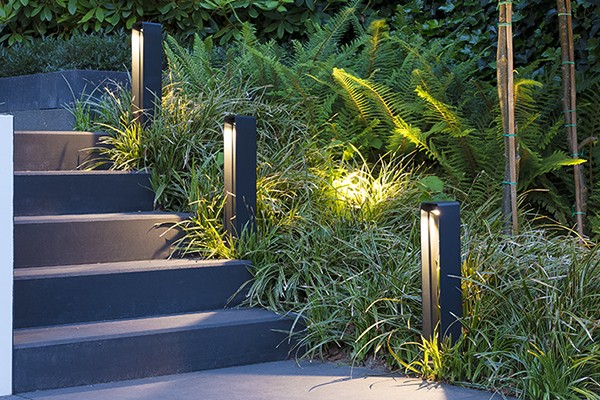 Die Wegeleuchte ACE von in-lite sorgt für eine dekorative Beleuchtung einer Außentreppe. Daneben ist ein beleuchtetes Beet mit Ziergräsern zu sehen.