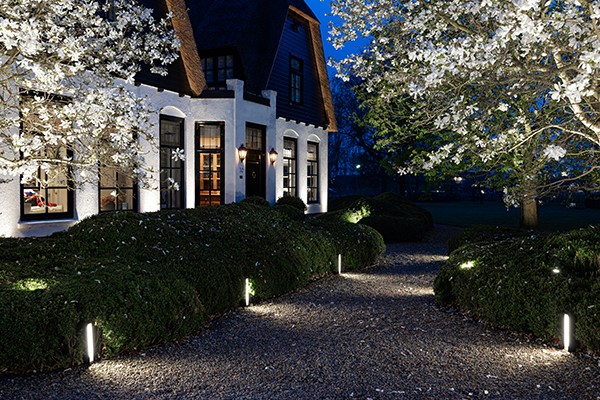 Ein Weg, der an einem Haus entlangführt, wird von ansprechender Gartenweg-Beleuchtung erhellt. Mehrere Stehleuchten markieren den Weg in regelmäßigen Abständen.